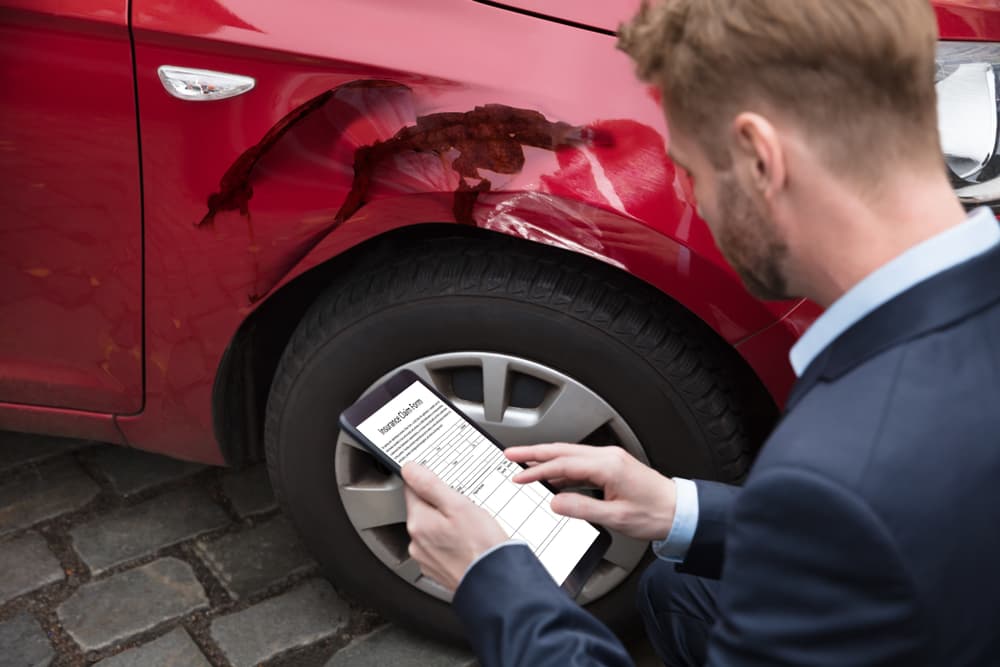 Man looking at a digital tablet screen displaying an insurance claim form near a car.